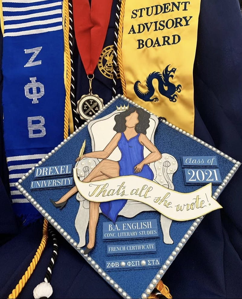 A commencement cap with the phrase "That's All She Wrote" and a seated woman on a chair.