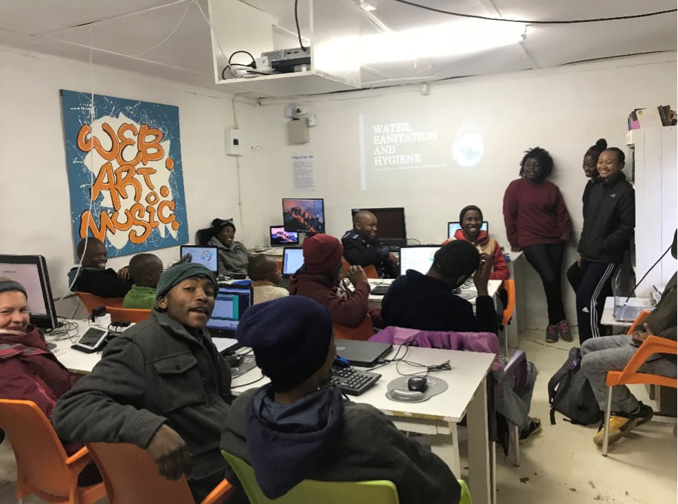 A classroom of people with three standing at the front next to a projection reading "Water Sanitation and Hygeine."