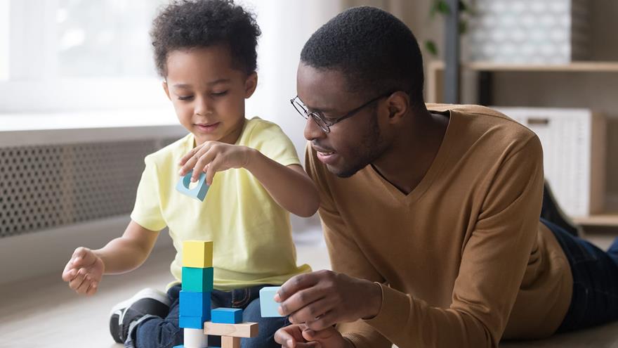 Dad lying on floor next to young son playing with colorful blocks