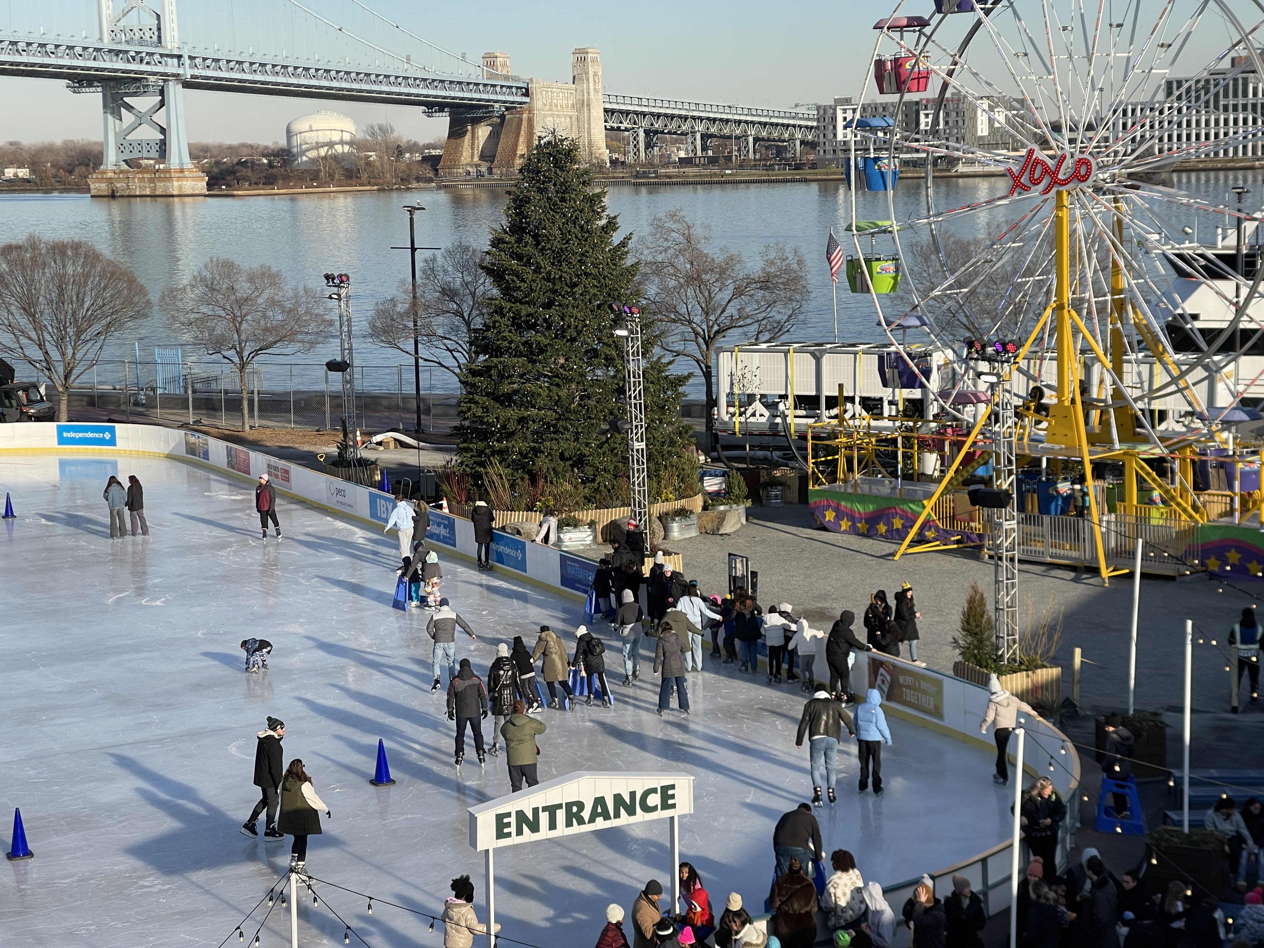 People ice skating outside on a rink next to a ferris wheel on the Delaware River.