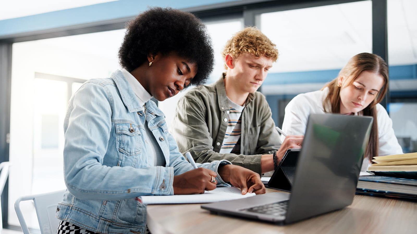 Three students sit in front of a lap top writing down notes
