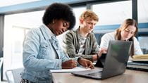 Three students sit in front of a lap top  writing down notes