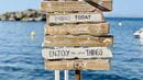 Wooden signs stacked on a beach with different sayings related to enjoying the moment.
