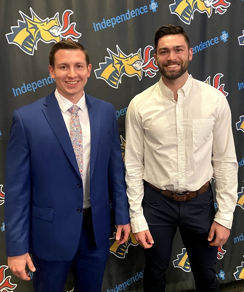 Two white males standing in front of a banner with the Drexel Dragon and the Independence Blue Cross logos