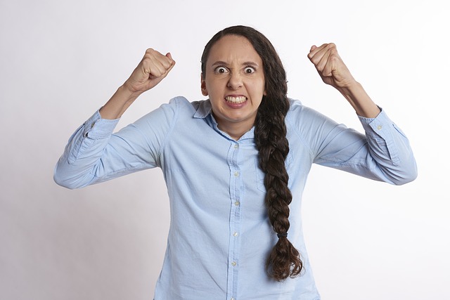Woman with a long thick braid of dark hair, wearing a light blue, long-sleeved button-down shirt, with her fists in the air looking very angry