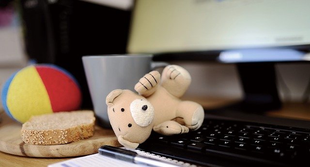 Image of a computer in a home office with a teddy bear sitting on the laptop.