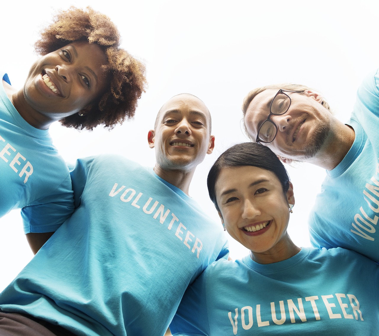 Image of 4 diverse people forming a circle around the camera, as in huddled - all wearing a light blue t-shirt that says "Volunteer" on the front.